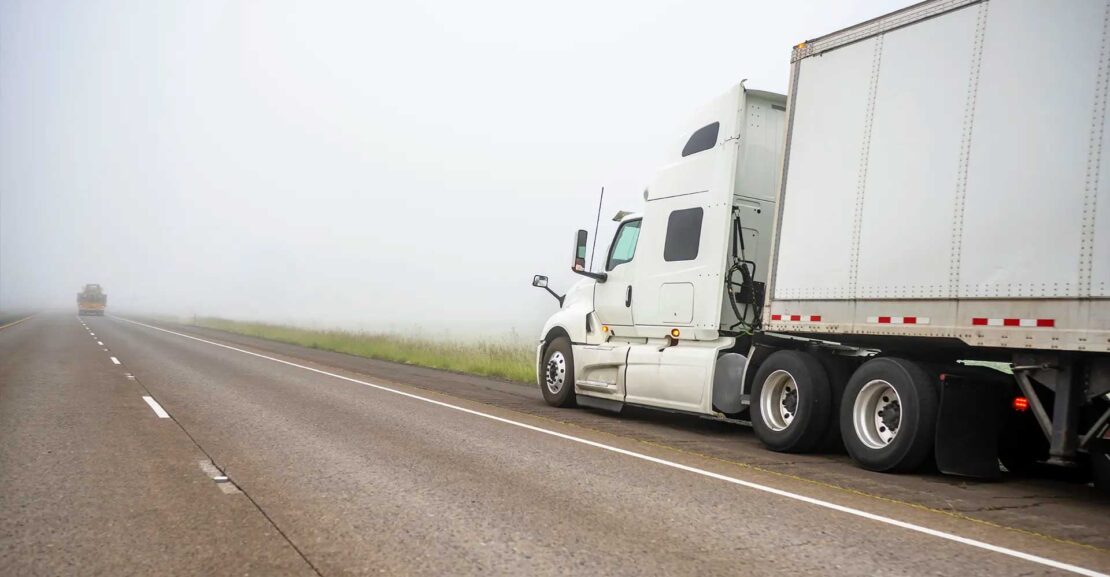 A semi-truck and trailer parked on a foggy highway shoulder waiting for roadside assistance